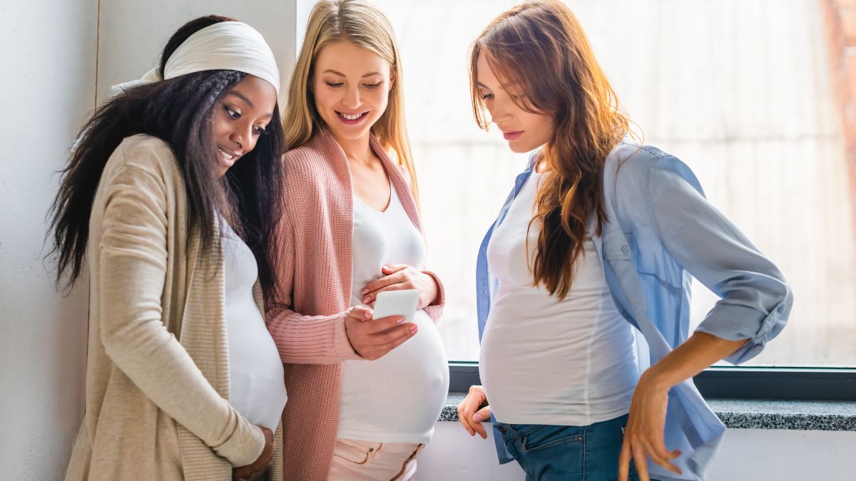 Three pregnant women discussing their surrogacy journey while looking at a cell phone during a family gathering.